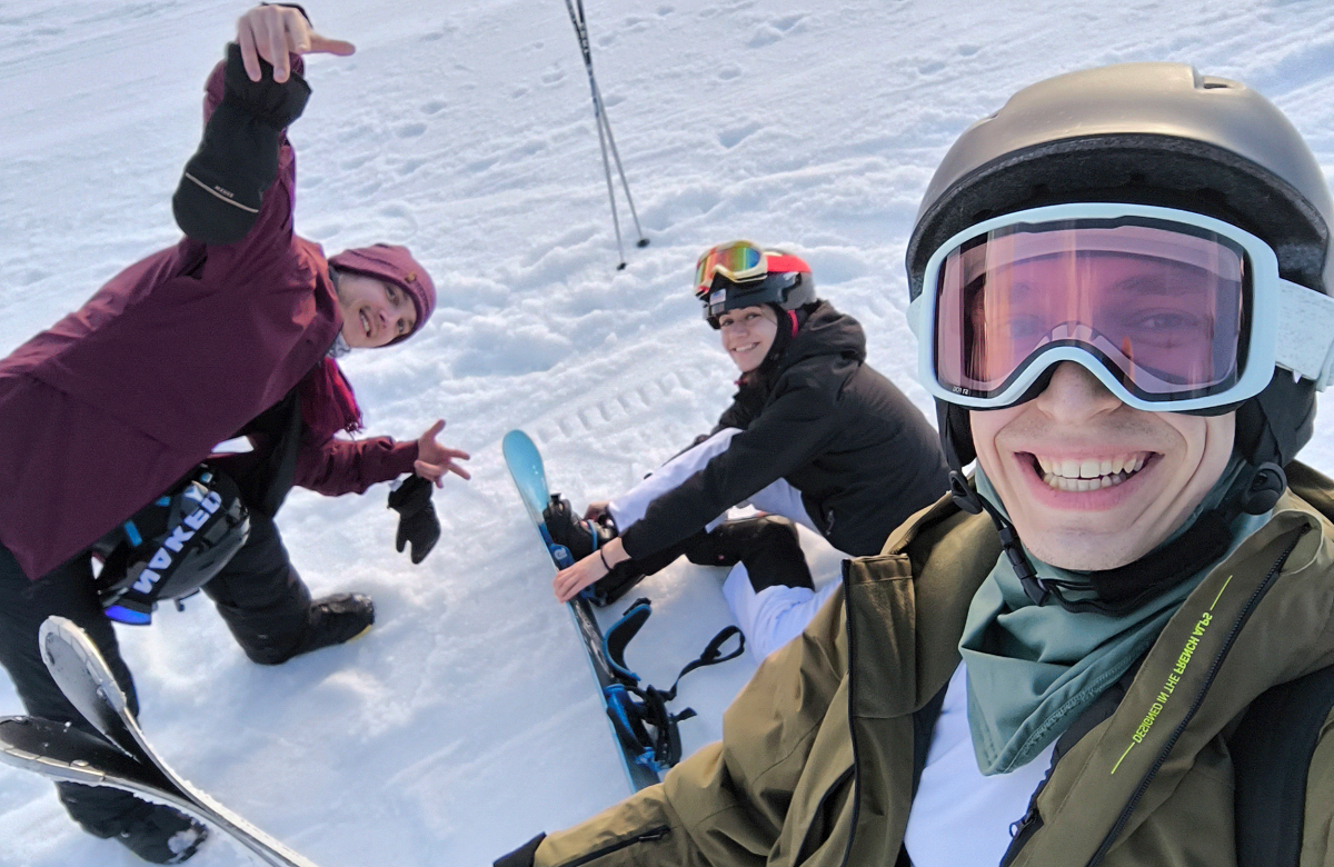 A group of friends smiling and posing for a selfie in their ski gear on a snowy mountain