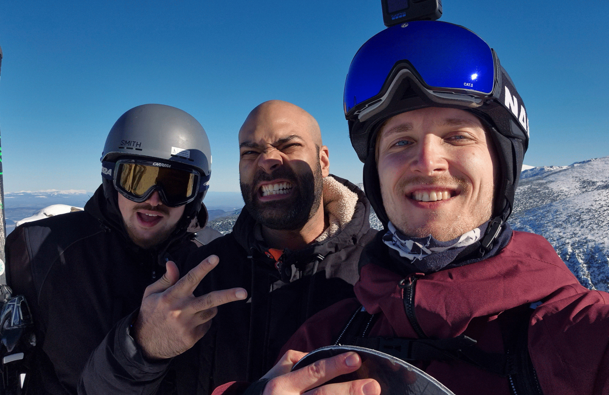 A group of friends in ski helmets and winter gear, posing for a selfie with mountains in the background